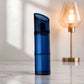 A translucent dark blue cosmetic pump bottle standing upright on a light marble surface, with a softly blurred warm-toned table lamp in the background.
