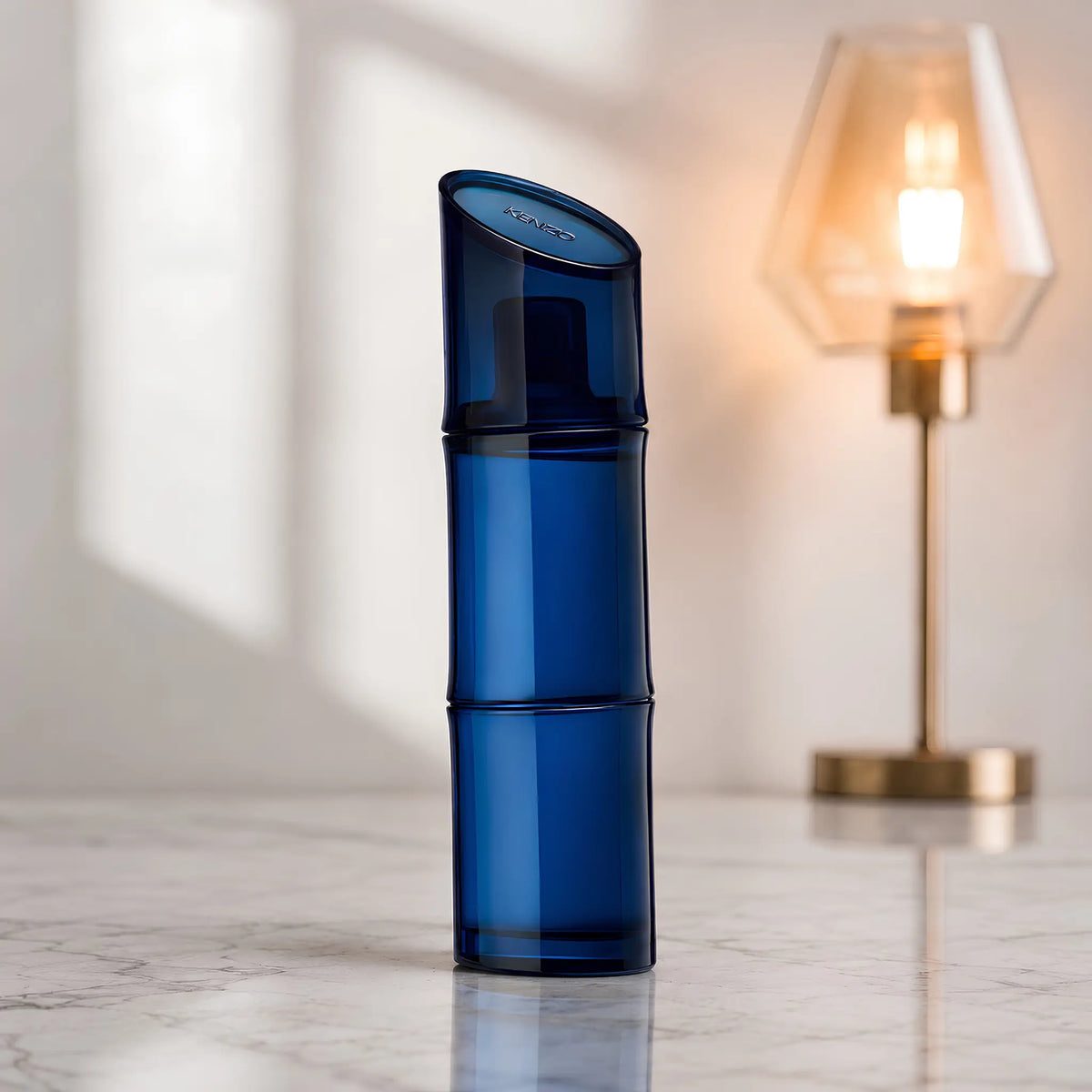 A translucent dark blue cosmetic pump bottle standing upright on a light marble surface, with a softly blurred warm-toned table lamp in the background.